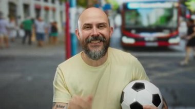 Sakallı, sakallı, elinde futbol topu olan, otobüslü ve arka planda yürüyen bir adam..