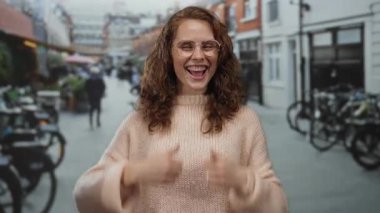 Şehir caddesinde gözlüklü, gülen bir kadın açık hava kentsel ortamının tadını çıkarıyor. Arka planda bisikletler ve binalar var..