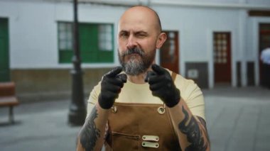 Sakallı ve dövmeli bir adam önlük takıyor ve eldiven takıyor. Arka planda bulanık binalar olan şehir caddesindeki kameraya bakıyor..