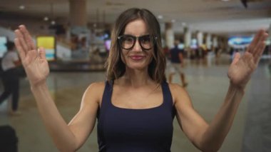 Genç bir kadın kalabalık havaalanı terminalinin içinde avuçlarını tutarken duruşu ve doğrudan bakışını koruyor; kendine güven.