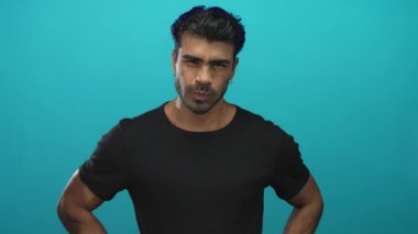 Siyah tişörtlü genç adam stüdyoda kameraya ciddi bir ifade ve dik duruşla işaret ediyor. Kararlılık..