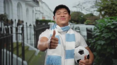 Sokakta gülümseyen ve elinde başparmağıyla futbol topu tutan atkı takan genç bir adam açık hava sporları coşkusu ve şehir yaşam tarzını öneriyor..