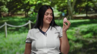 Güneşli park ortamında güzellik süngeri tutan bir kadın, günlük beyaz gömlekli bir kadın, yemyeşil arka planda neşeyle gülümseyen, doğal güzellik ve rahatlamayı vurgulayan.