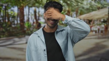 Genç Hispanik adam açık kafenin yanındaki güneşli yeşil parkta tropikal yeşillikler arasında sağ eliyle gözlerini kapar; neşe dolu neşe.