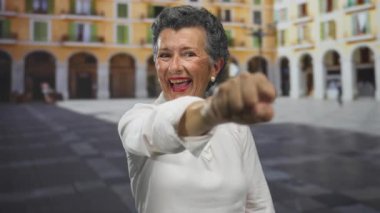 Gri saçlı yaşlı bir kadın arka planda canlı bir mimarisi olan eski bir şehir meydanında neşeli bir şekilde yumruğunu uzatıyor..