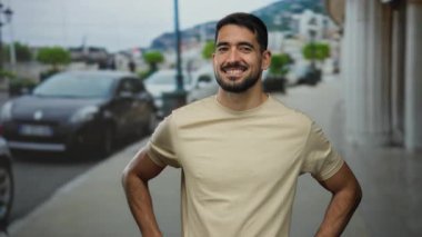 Kentsel bir sokak ortamında güvenle gülümseyen genç bir adam, rahat bir poz sergiliyor arabaların ve binaların arka planına karşı, modern ve canlı bir şehir yaşamını somutlaştırıyor..