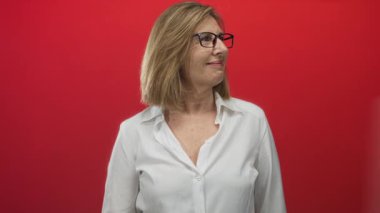 Kırmızı stüdyoda beyaz gömlekli ve gözlüklü bir kadın başparmağını omzuna uzatırken gülümsüyor..