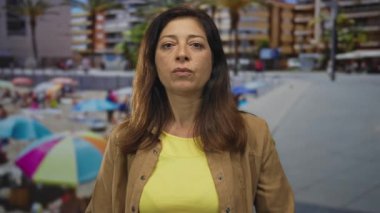 Sarı gömlekli ve kahverengi ceketli orta yaşlı İspanyol kadın sokakta güneş ışığı altında duruyor..