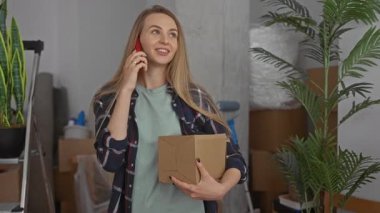 Yeni bir evde telefonla konuşan bir kadın. Etrafı bitkilerle ve kutularla çevrili. İçerideki rahat ortamında heyecan ve beklenti hissi yayıyor..