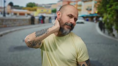 Sakallı ve dövmeli kel adam şehir sokağında duruyor. Arka planda bulanık binalar var. Yakışıklı ve açık hava havasını yansıtıyor..