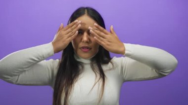 Uzun saçlı genç bir kadın canlı mor bir arka plan karşısında sevinçle el kol hareketi yapıyor beyaz bir kazak giyiyor, pozitiflik ve enerji taşıyor..