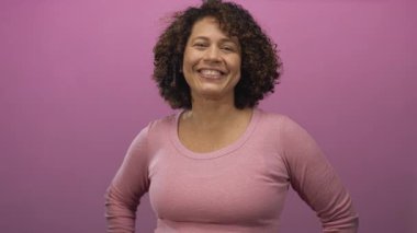 Pembe bluzlu İspanyol kadın, elleri kalçasında mor stüdyo duvarına yaslanmış kameraya bakıyor..