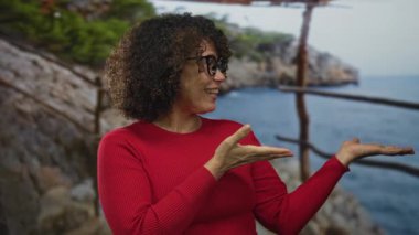 Gözlük takan kadın, kayalık sahile ve denize tepeden bakan tırabzanlar yaparak avuçlarını açık tutarak jestler yapıyor..