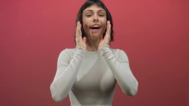 Beyaz kesim bluzlu bir kadın iki eliyle bir stüdyoda yanaklarına dokunuyor; mutluluk..