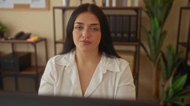 Ofiste parmaklarıyla hayır işareti yapan bir kadın, bir bilgisayarın önünde oturuyor, raflar, bitkiler ve klasörlerle çevrili, profesyonel bir ortamda beyaz gömlek giyiyor..