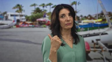 Orta yaşlı İspanyol kadın, arkasında yelkenli ve palmiye ağaçları olan bir limanı işaret ediyor..