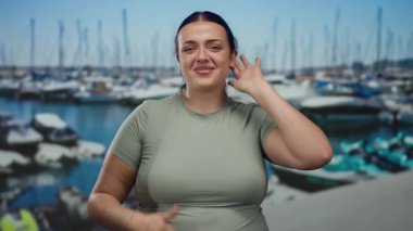 Arka planda tekneleri olan, deniz kıyısında gülümseyen genç ve cüsseli bir kadın, açık hava ve sakin atmosferin tadını çıkarıyor, pozitif ve kendinden emin bir tavır sergiliyor..