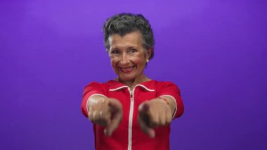 Kadın kendine güvenerek kameraya doğru gülümsüyor, canlı mor bir arka plan karşısında duruyor, renkli kırmızı bir elbiseyle pozitif enerji yayıyor..