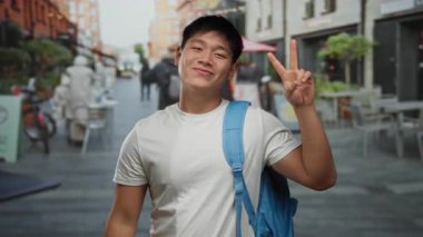 Genç adam gülümsüyor ve açık bir kahve dükkanının terasında barış işareti yapıyor. Kalabalık bir şehir restoranında rahat bir atmosferi var..