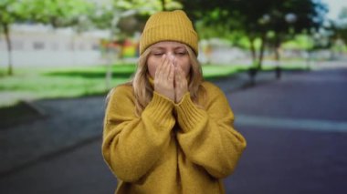 Sarı süveter içinde titreyen ve açık hava parkında örgü ören bir kadın soğuğu ifade ediyor, arka planda ağaçlar ve parlak gün ışığı var..