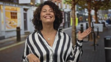 Kıvırcık saçlı, çizgili bluz giyen bir kadın, mağaza vitrinleriyle kaplı bir sokakta avuç içini açmak için işaret ediyor..