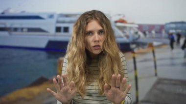 Sıradan sahnelerde kalabalık bir limanda elini kaldıran bir kadın görülüyor. Sarı saçları, feribot iskelesinde çizgili bir süveteri çerçeveliyor..