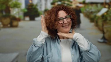 İspanyol kadın hareketli bir şehir caddesinde neşeli jestler yapıyor, canlı bitkiler ve hafif ışıklandırmayla çerçevelenmiş, canlı bir açık şehir anını yakalıyor..