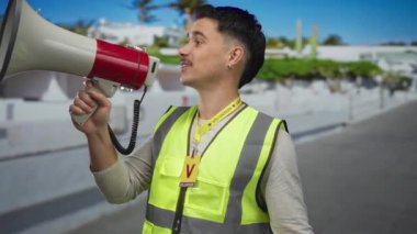 Şehir caddesinde megafonu gönüllü olarak kullanan yansıtıcı yelek giyen genç adam, şehir manzarası ile çevrili, coşkuyla halkı meşgul ediyor..