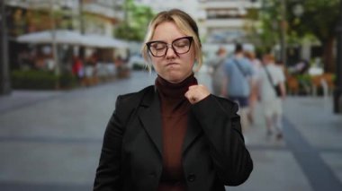 Gözlüklü, sarı saçlı, sıradan giyinmiş bir kadın hareketli bir şehir caddesinde duruyor. Yumruğunu sıkarak ve odaklanmış bir bakışla kararlılığını gösteriyor..