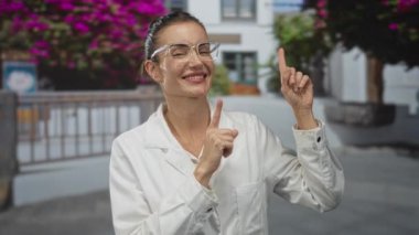 Beyaz laboratuvar önlüğü ve gözlüklü kadın bilim adamı, binanın dışındaki mor çiçeklerle objeyi işaret ediyor..