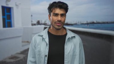 Genç adam iki eliyle yanakları bastırıyor ve bulutlu gökyüzünün altında deniz kenarında beyaz bir duvarın yanında dudaklarını büzüştürüyor..