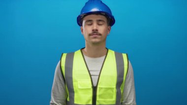 Kask takan ve yansıtıcı yelek giyen İspanyol bir adam mavi arka plan duvarına karşı gözlerini kapatarak rahatlama hissini iletiyor ve sakin bir ortama odaklanıyor..