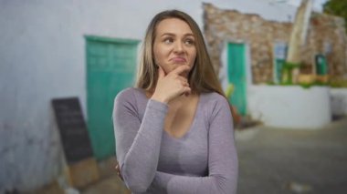 Kırsal yeşil kapının ve taş duvarın önünde kollarını kavuşturarak dışarı çıkmayı düşünen düşünceli bir ifade ve günlük giysiler sergileyen bir kadın..