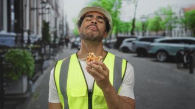 Meşgul şehir caddesinde altın waffle yiyen adam sarı yelek ve mühendis şapkası giyiyor; memnuniyet.