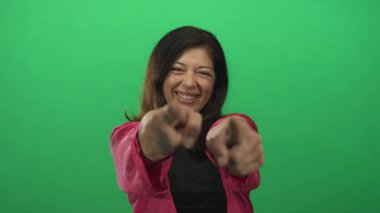 Stüdyoda pembe ceketli, gülümseyen ve kamerayı yeşil ekrana gösteren bir kadın..