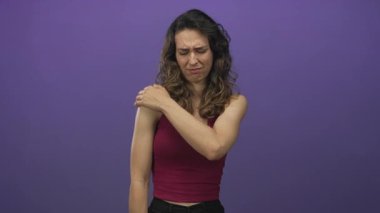 Kırmızı kolsuz bluz giyen genç İspanyol kadın mor stüdyo ortamında ürkerken çıplak omzunu tutuyor..