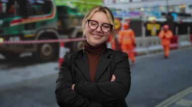 Gözlüklü ve siyah ceketli sarışın kadın işçiler ve engellerle çevrili yoğun bir şantiyede kollarını kavuşturarak profesyonellik ve yetkinliği yansıtıyor..