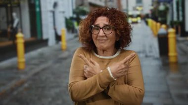 Orta yaşlı, kıvırcık saçlı İspanyol kadın şehir sokaklarında kararsız bir şekilde iki tarafı da işaret ediyor, açık hava ortamında belirsizliğini dile getiriyor..