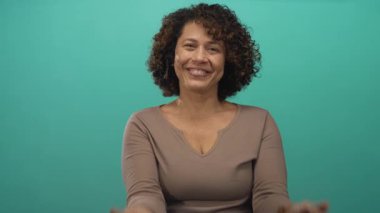 Kıvırcık saçlı bir kadın, el ele tutuşurken kalp hareketi yaparken ve turkuaz bir stüdyoda gülümserken; sevgi mutluluğu sıcak neşe.