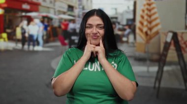Çevrede canlı insanlar ve kafelerle dolu hareketli şehir ortamında yeşil tişörtlü, kıvırcık kadın gönüllü gülümsüyor.