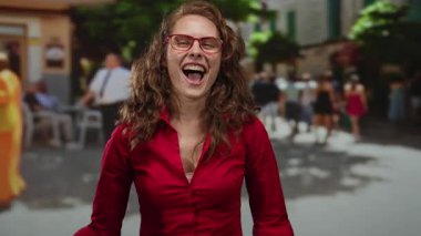 Kırmızı gömlekli, gözlüklü bir kadın, arka planda bulanık yayalarla dolup taşan bir şehir caddesinde kendini gösterirken kendine güvenerek gülümsüyor..