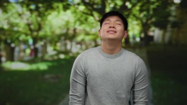Gri kazaklı genç adam yemyeşil ağaçlar ve ışık saçan huzurlu bir parkın keyfini çıkarıyor.
