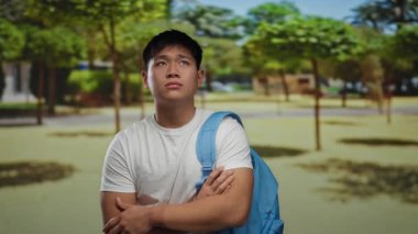 Genç adam güneşli bir parkta kollarını kavuşturmuş, tişört ve sırt çantası giymiş, açık havada bir doğal ortamda iç gözlem anı yakalıyor..