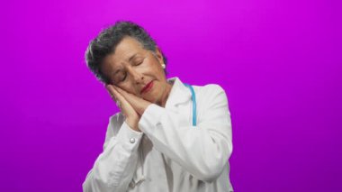 Steteskoplu kıdemli kadın doktor canlı pembe arka plana karşı uyku jesti gösteriyor. Bu da yorgunluğu ya da tıp mesleğinde dinlenmeyi gösteriyor..