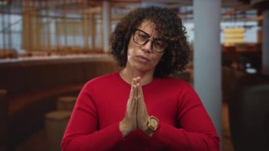 Kadın restoranda kırmızı kazak ve gözlük takarak el ele tutuşuyor, gözleri dua pozisyonunda kapalı; huzur umudu.