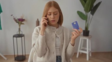 Oturma odasındaki genç sarışın kadın elinde kredi kartıyla konuşuyor. İç mekan dekoruyla ilgileniyormuş..