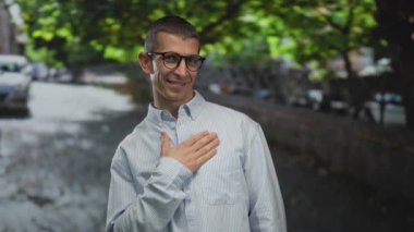 Ağaçlı, gözlüklü, çizgili tişörtlü, göğsünde elinle gülümseyen yeşil yaprakların altında dostça bir şehir atmosferi yaratan bir adam..