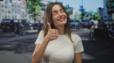 Tişört giyen kadın, işlek caddede baş parmak hareketi gösteriyor; kendine güven iyimserliği teşvik.