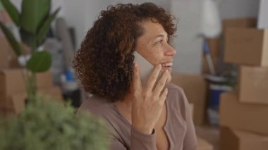 Bir binadaki yemyeşil bir saksı bitkisinin yanında kutuları taşıyan akıllı telefonu kulağına tutan kadın; memnuniyet.