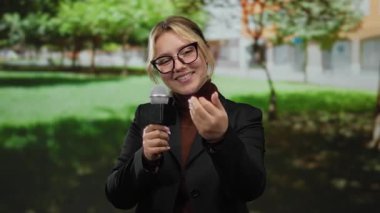 Parkta bir kadın muhabir elinde mikrofon ve işaret parmağıyla elini gözlük takıp güneşli bir gökyüzünün altında yeşillikle çevrili bir şekilde buraya geliyor.